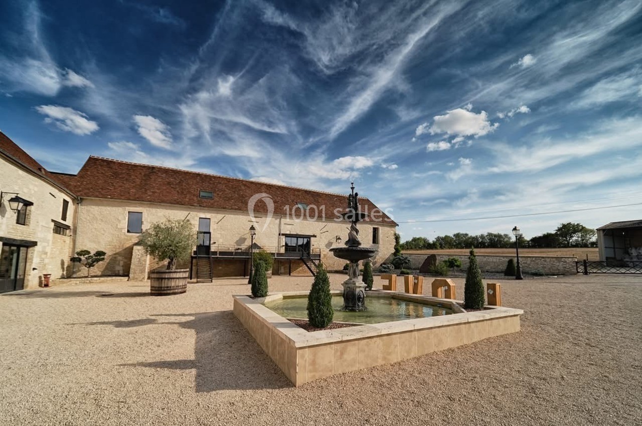 Cour pavée avec une fontaine centrale, entourée de bâtiments en pierre sous un ciel bleu parsemé de nuages.