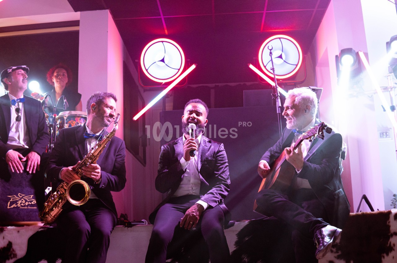 Un chanteur accompagné de musiciens jouant du saxophone et de la guitare sur une scène éclairée.