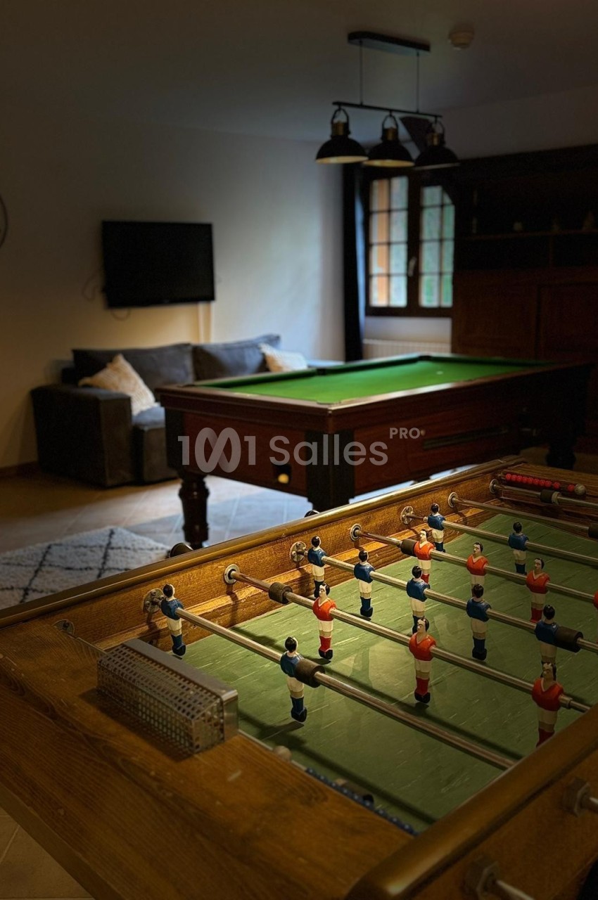 Baby-foot au premier plan avec un billard en arrière-plan dans une pièce aménagée avec canapé et télévision.