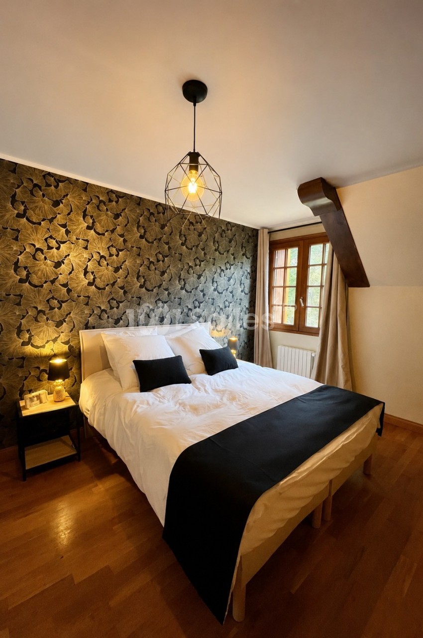 Chambre lumineuse avec lit double, linge blanc, coussins noirs, mur à motif floral sombre et fenêtre en bois.