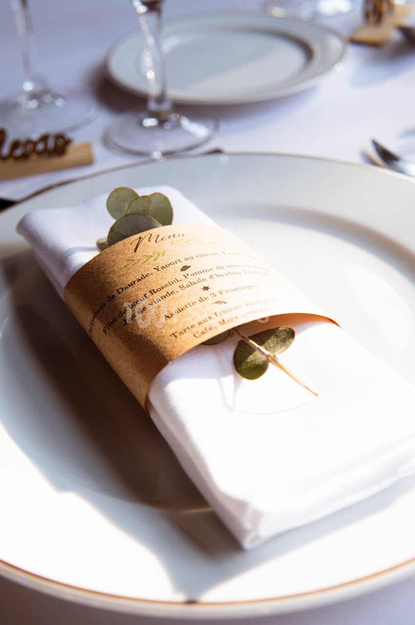 Serviette blanche pliée avec un menu enroulé et une branche d'eucalyptus, posée sur une assiette blanche.