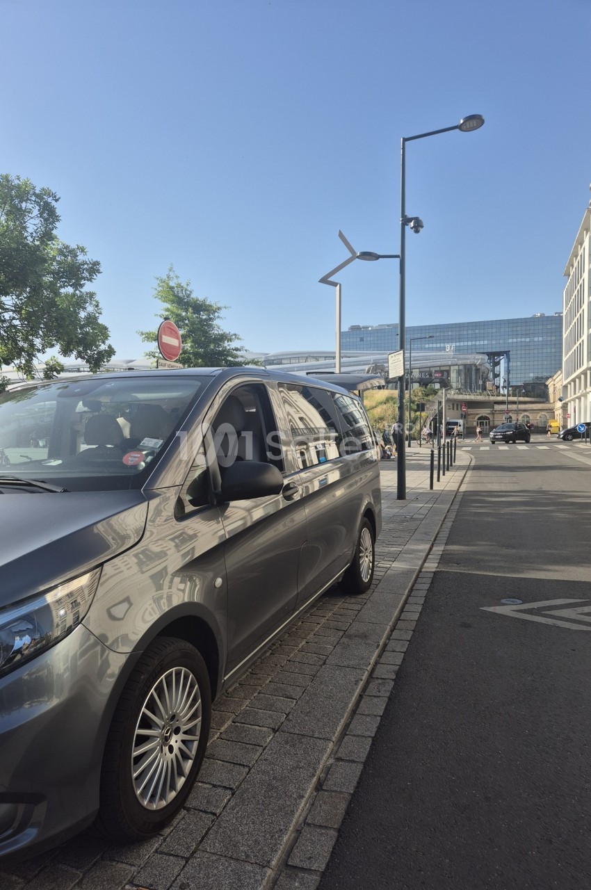 Un van gris stationné le long d'un trottoir dans une rue urbaine avec des bâtiments modernes en arrière-plan.