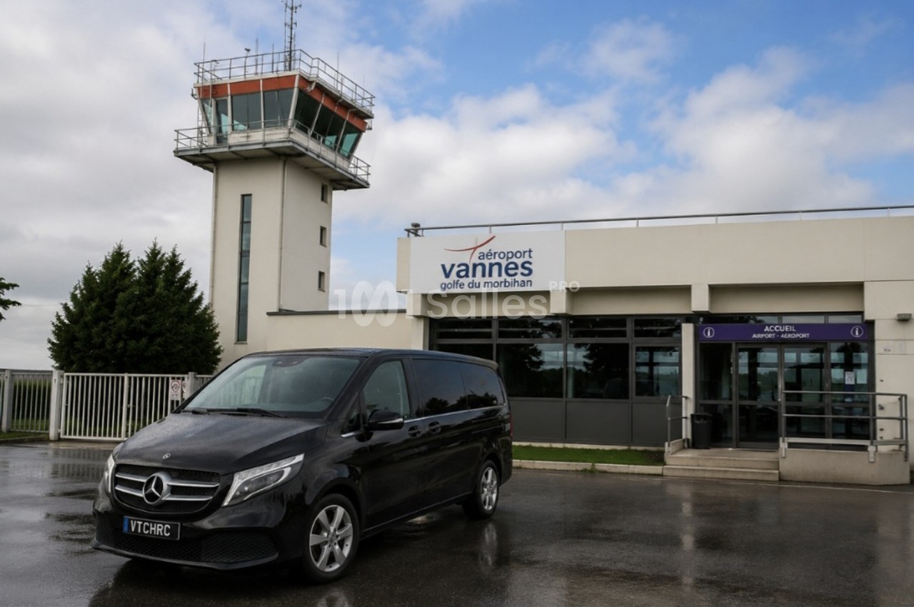 Minibus noir stationné devant l'entrée de l'aéroport de Vannes avec une tour de contrôle en arrière-plan.