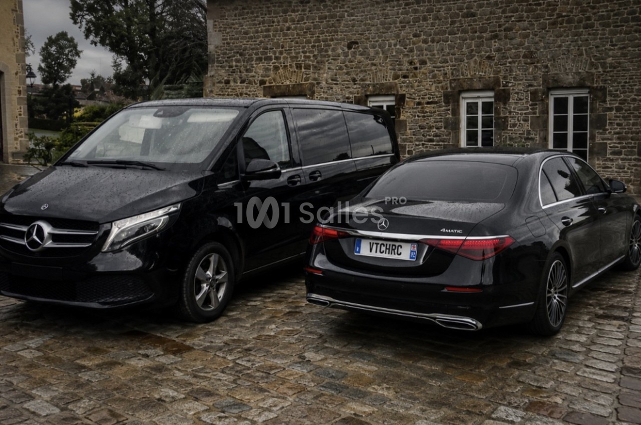 Deux véhicules Mercedes noirs stationnés sur un sol pavé devant un bâtiment en pierre sous un ciel nuageux.