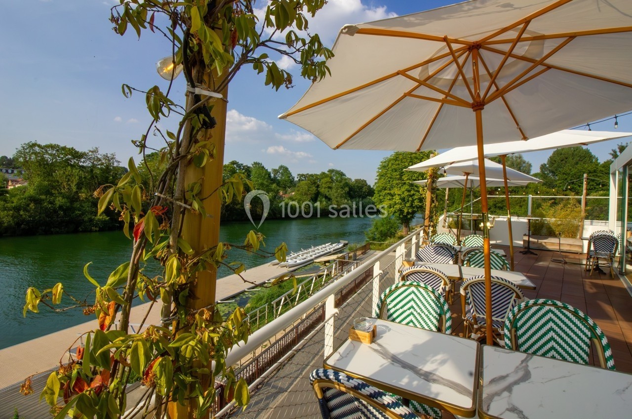 Terrasse ensoleillée avec tables, chaises et parasols, surplombant une rivière entourée de verdure.