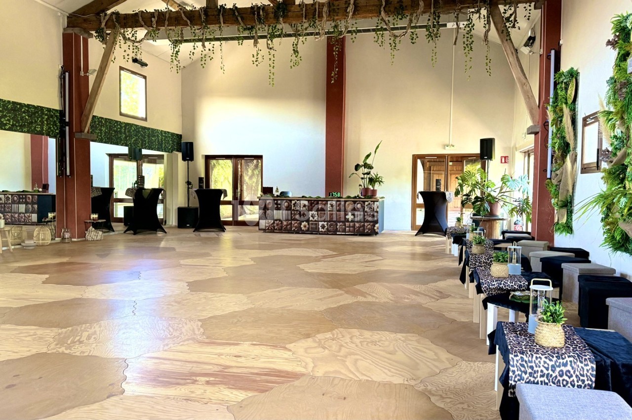Salle spacieuse avec parquet clair, poutres apparentes, décoration végétale et tables basses alignées.