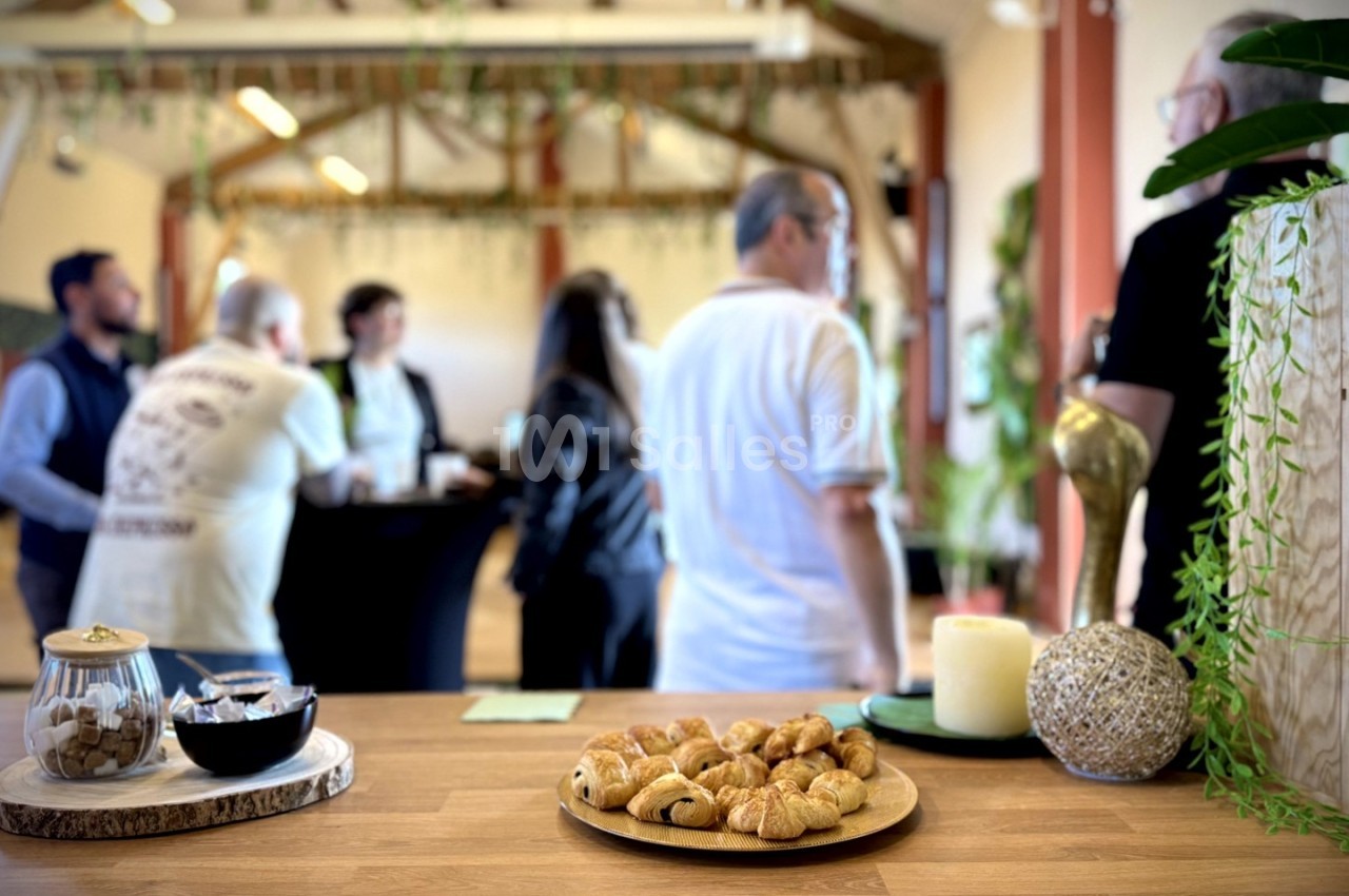 Des viennoiseries et biscuits sur une table en bois, avec des personnes discutant en arrière-plan dans un espace lumineux.
