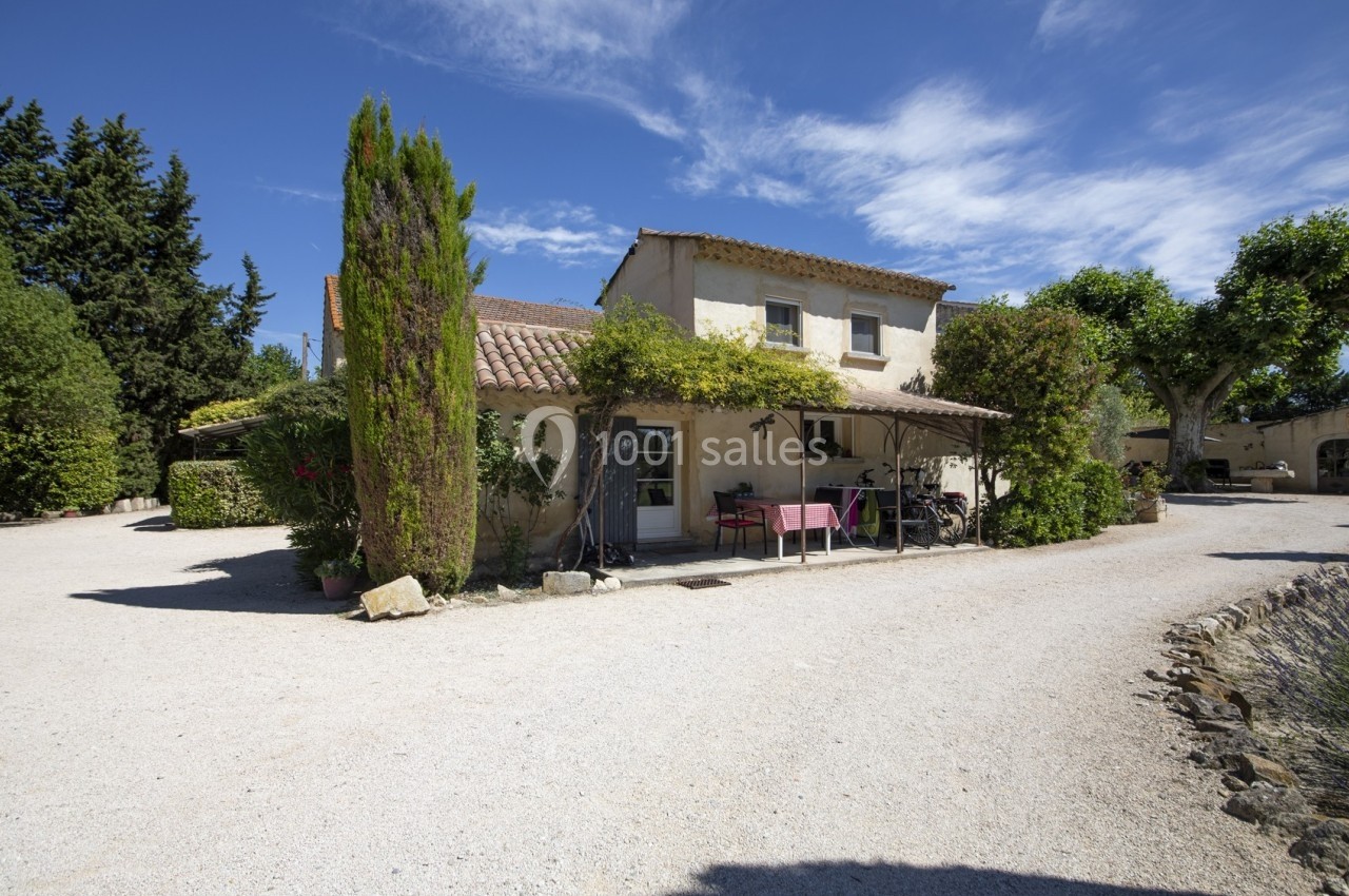 Maison en pierre avec toit en tuiles, terrasse ombragée et végétation environnante, sous un ciel bleu clair.