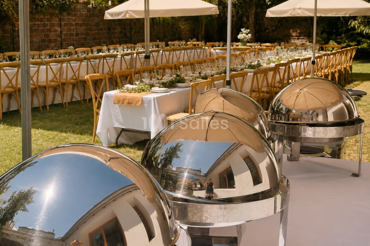 Espace de réception en jardin avec tables dressées, chaises en bois, parasols blancs et cloche chauffante au premier plan.