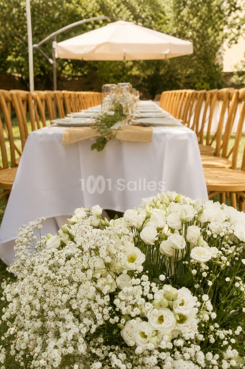 Jardin avec table ronde dressée pour réception, chaises dorées et parasol blanc, fleurs blanches en premier plan.