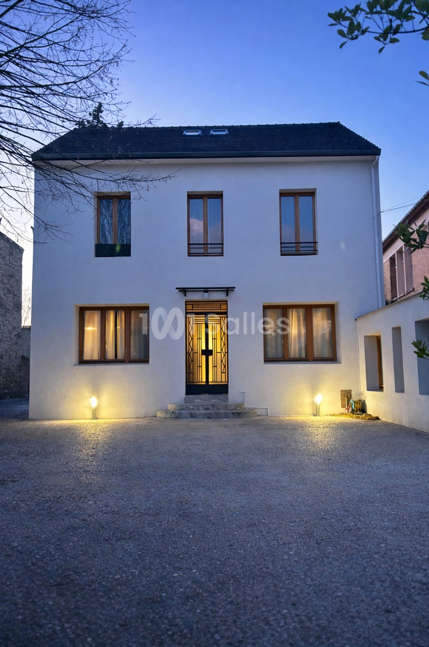 Façade blanche contemporaine avec toit ardoisé, éclairée de nuit, entrée centrale et cour pavée.