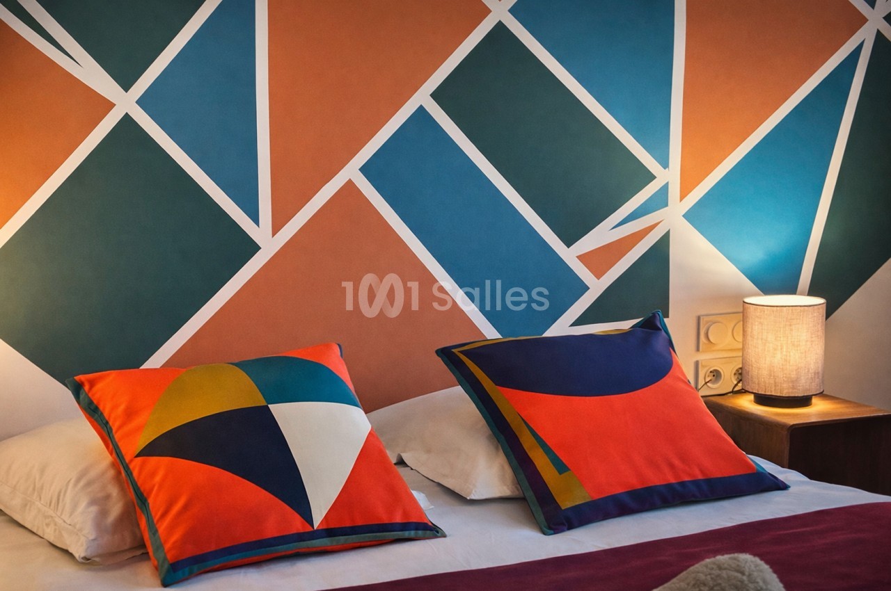 Chambre avec lit aux coussins géométriques multicolores, tête de lit en losanges bleus, oranges et verts, lampe de chevet allumée.