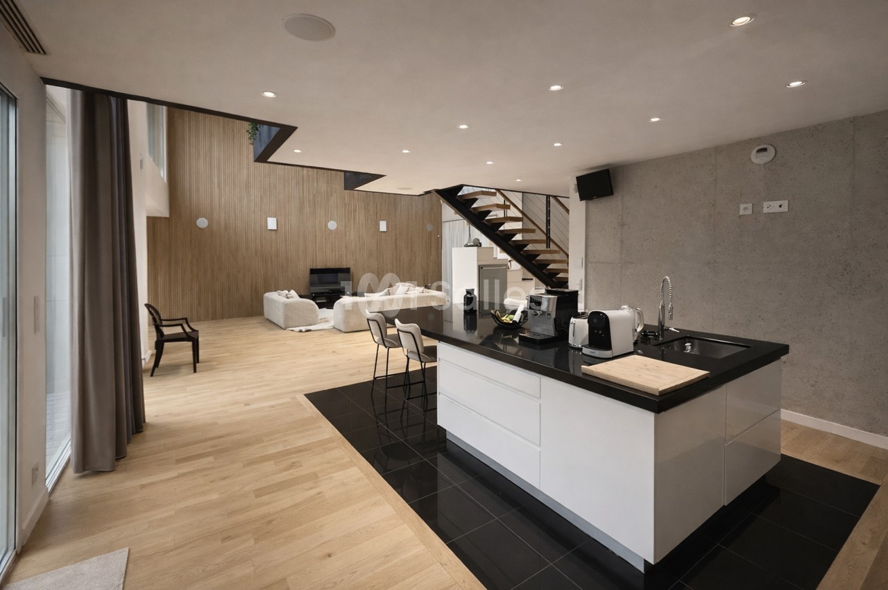 Cuisine ouverte avec îlot blanc et plan de travail noir, séjour avec canapés beige et escalier bois en arrière-plan.