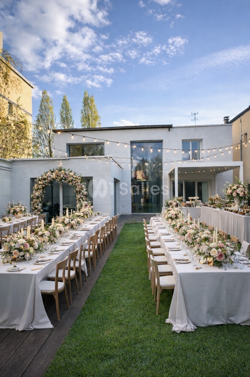 Cour intérieure aménagée pour un repas de mariage avec deux tables longues, chaises en bois, fleurs blanches et roses, guirlandes lumineuses.