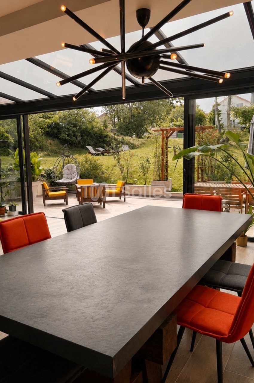Salle intérieure avec table grise et chaises rouges, baie vitrée ouverte sur jardin, plafond moderne avec lustre noir.
