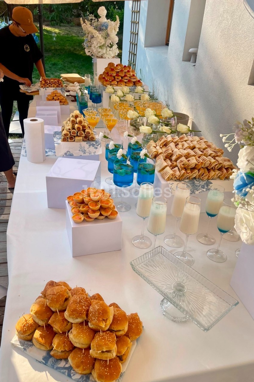 Table de réception dressée avec petits pains, pâtisseries, verres de jus orange et bleu, fleurs blanches, en terrasse ensoleillée.