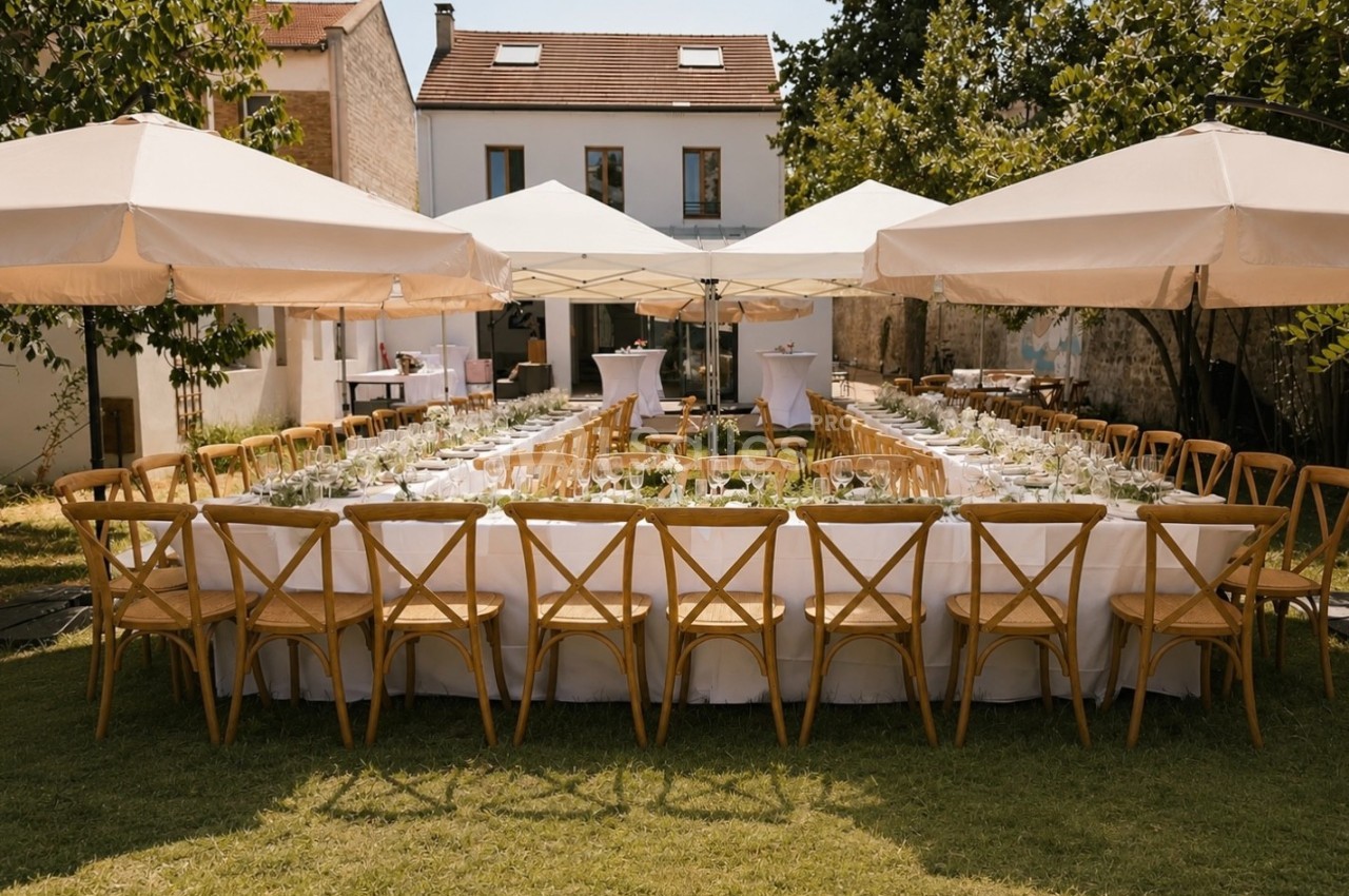 Table rectangulaire dressée en banquet dans un jardin avec chaises bistrot bois, nappes blanches et parasols beige.