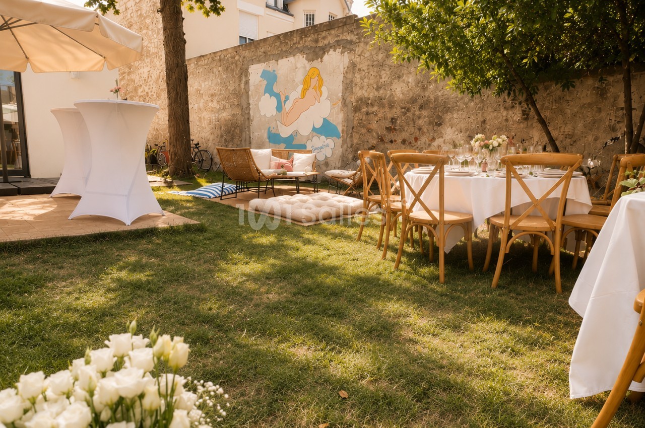 Jardin intérieur dressé pour réception avec tables rondes nappes blanches, chaises bois et mur en pierre orné d'une fresque.