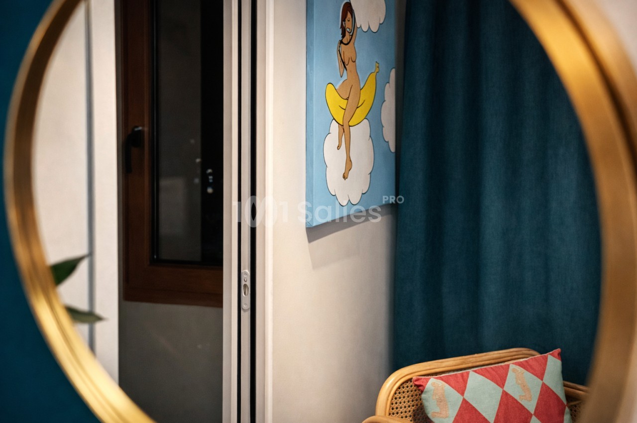 Couloir intérieur avec murs bleu canard, affiche vintage colorée et siège en rotin à motifs géométriques.
