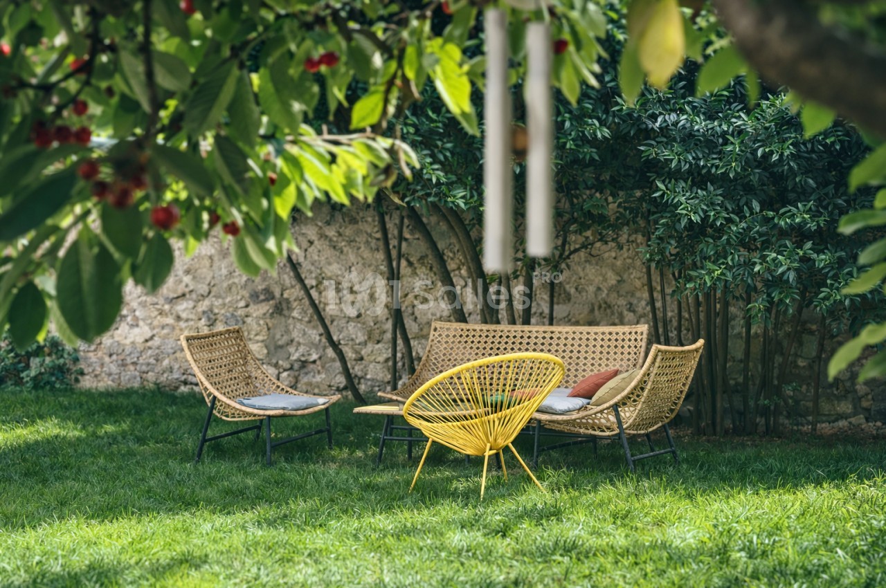 Salon de jardin avec trois chaises en rotin et table jaune sur pelouse, mur de pierre et végétation.