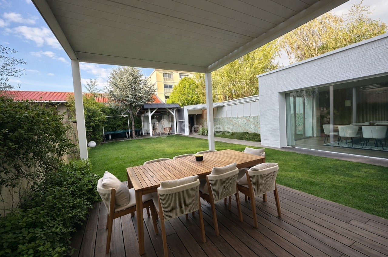 Terrasse couverte avec table en bois et chaises beige - jardin herbeux et bâtiment blanc en arrière-plan.