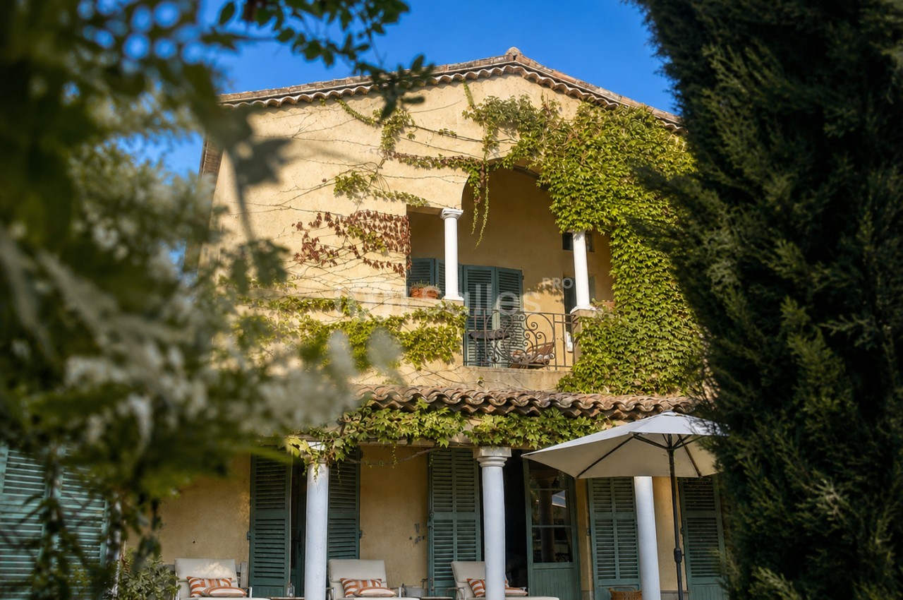 Façade en pierre ocre d'une maison provençale avec colonnes blanches, terrasse ombragée et volets verts.