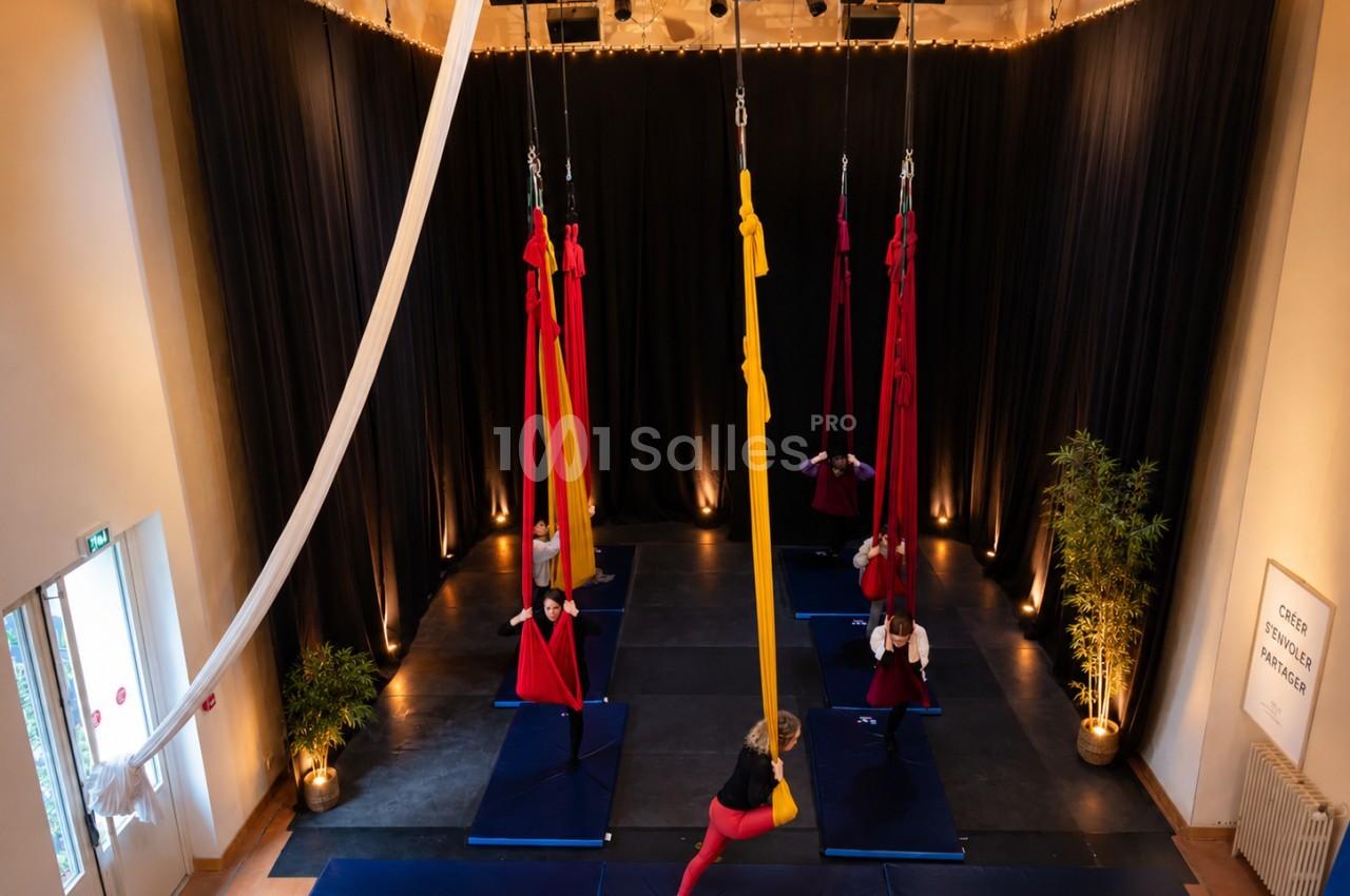 Personnes pratiquant le yoga aérien dans une salle avec des tissus suspendus, éclairage chaleureux et tapis au sol.