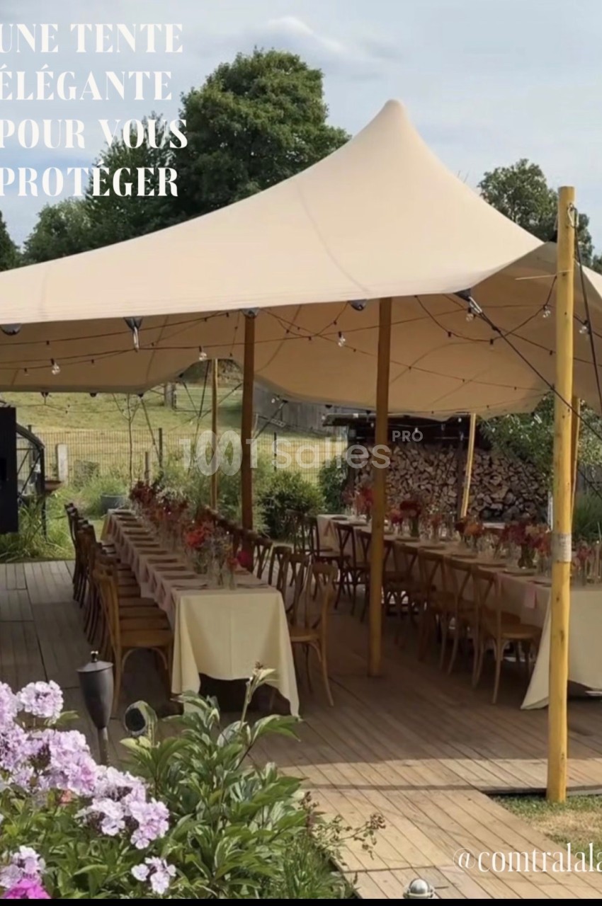 Tente beige installée en extérieur, couvrant des tables et chaises en bois, entourée de verdure et de fleurs.