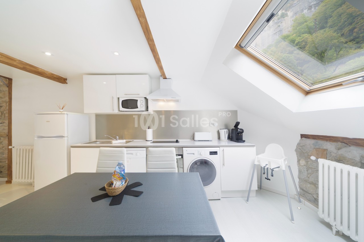 Cuisine moderne sous combles avec table, électroménagers, chaise haute et fenêtre de toit offrant une vue sur la nature.