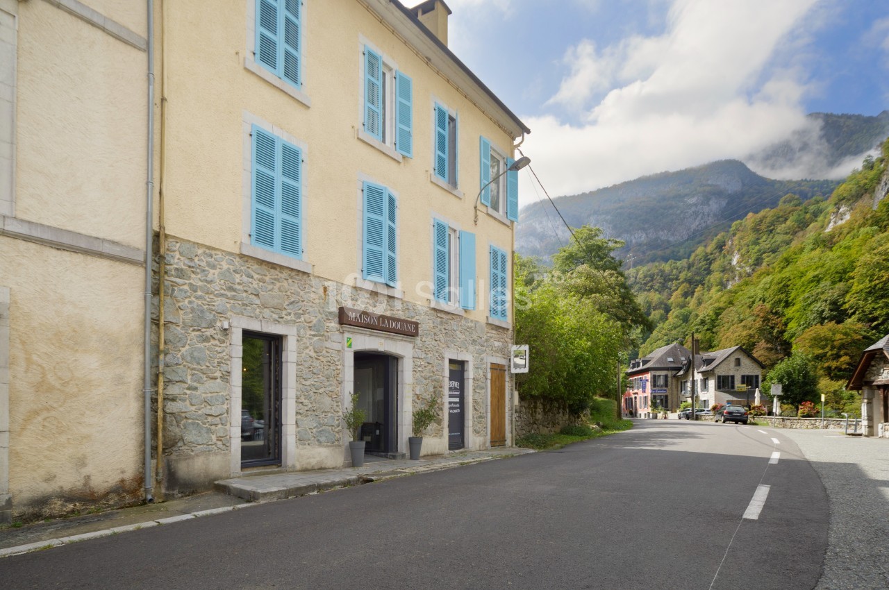 Façade d'un bâtiment en pierre avec volets bleus, situé dans un village entouré de montagnes et de végétation.