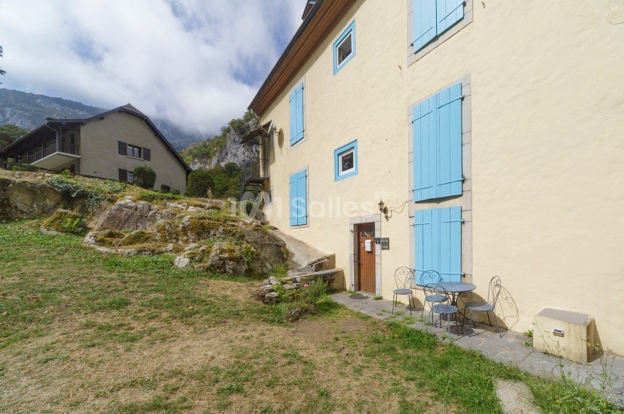 Façade d'une maison beige avec volets bleus, table et chaises en fer forgé sur une terrasse en pierre, jardin en pente.