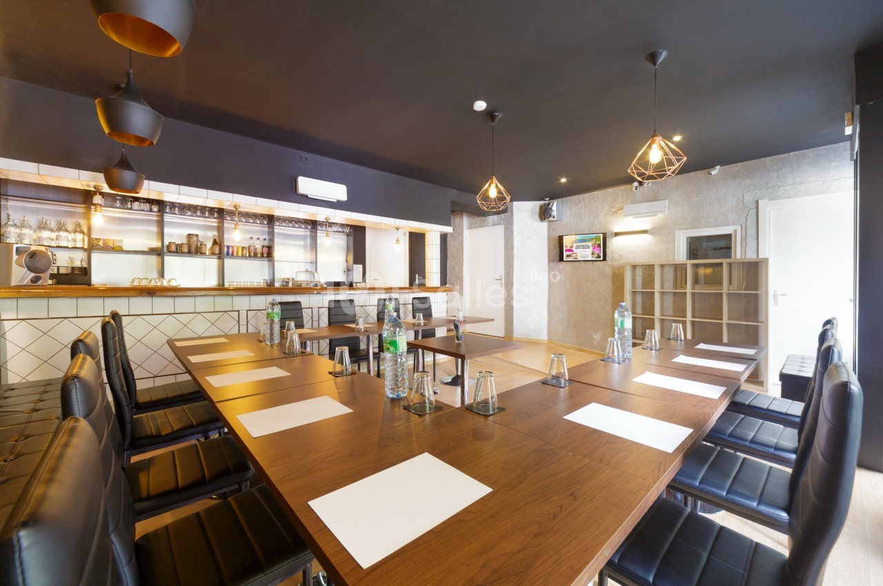 Salle de réunion moderne avec grande table en bois, chaises noires, éclairage suspendu et bar en arrière-plan.