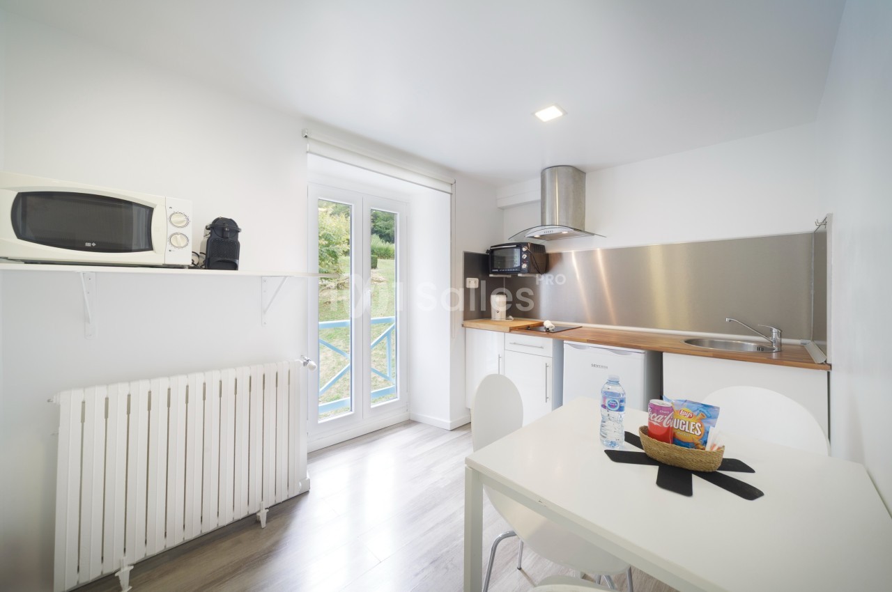 Cuisine lumineuse avec micro-ondes, cafetière, évier, table et chaises, donnant sur une fenêtre avec vue extérieure.
