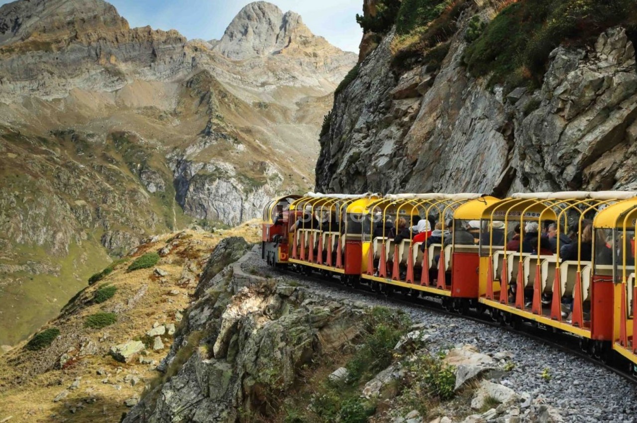 Train touristique rouge et jaune circulant sur une voie étroite, longeant une montagne rocheuse dans un paysage alpin.