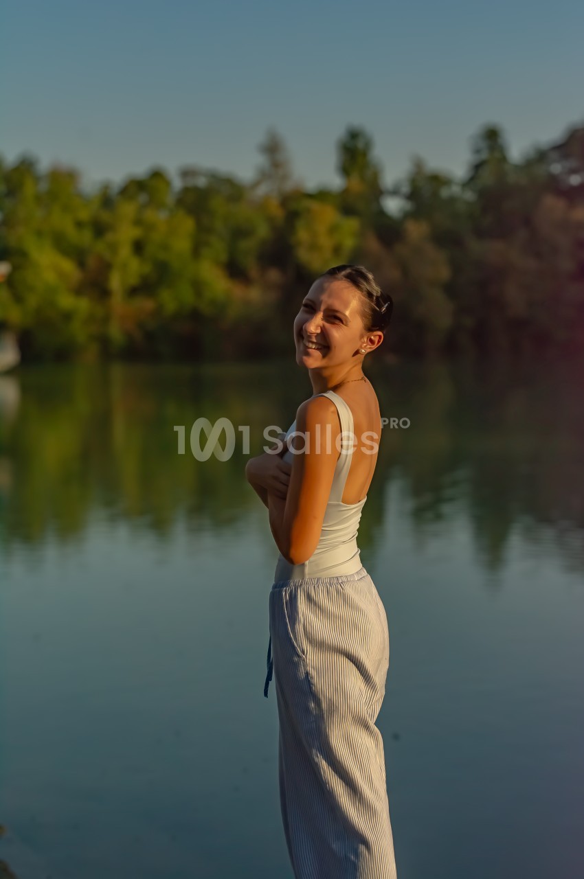 Femme souriante debout près d'un lac au coucher du soleil, entourée de végétation.