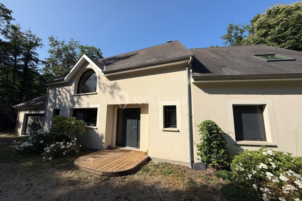 Maison moderne avec façade beige, toit en ardoise, terrasse en bois et jardin arboré sous un ciel dégagé.