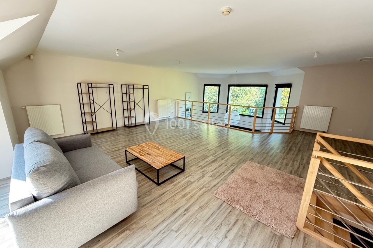 Salon spacieux avec parquet clair, canapé gris, table basse en bois, étagères et grandes fenêtres donnant sur la verdure.