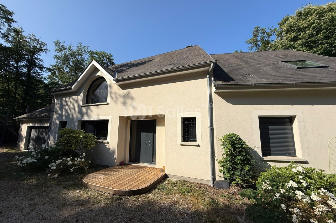 Façade d'une maison moderne avec toit en ardoise, grande fenêtre cintrée et terrasse en bois, entourée de végétation.