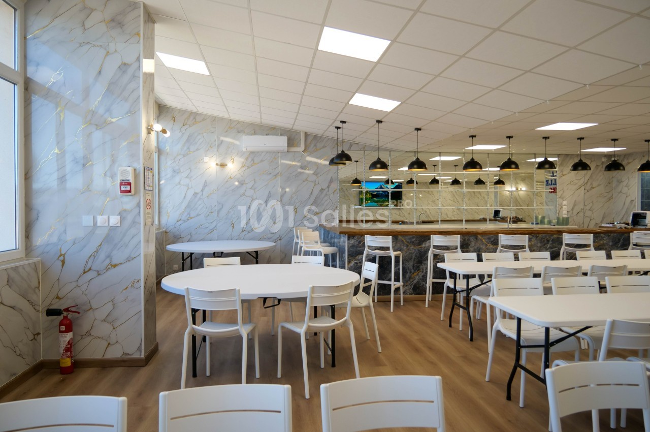 Salle lumineuse avec tables et chaises blanches, sol en bois clair, murs effet marbre et comptoir avec suspensions noires.
