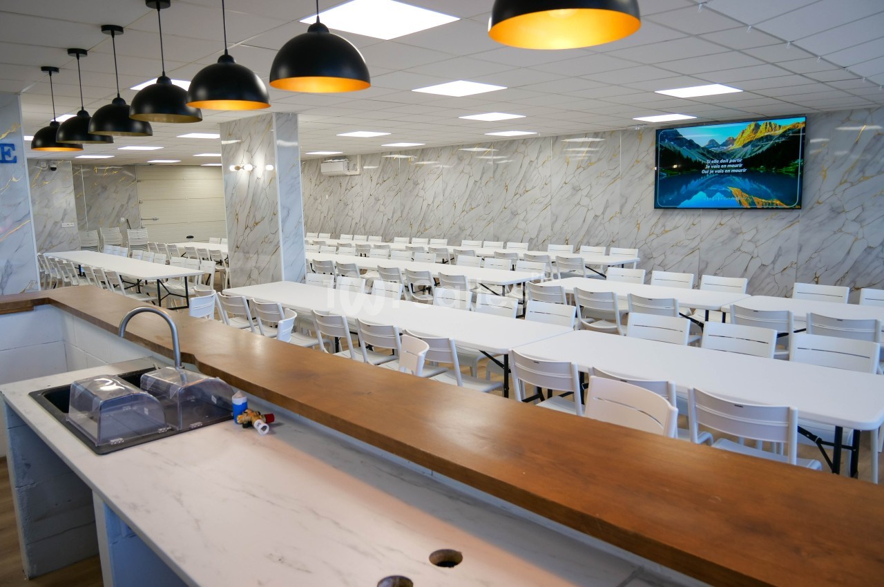 Salle à manger spacieuse avec tables blanches alignées, comptoir en bois au premier plan et écran mural affichant un paysage.