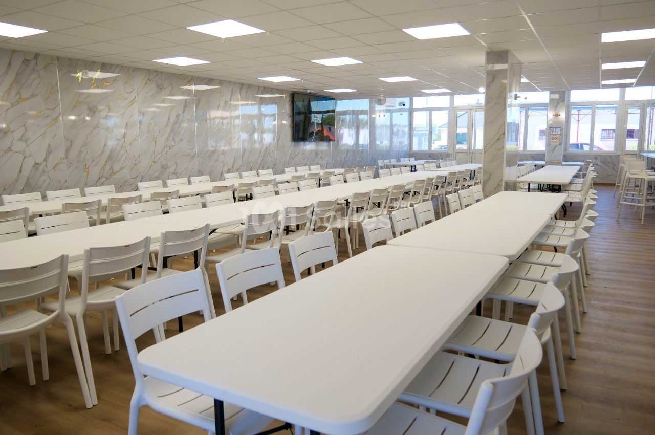 Salle lumineuse avec de longues tables blanches alignées et des chaises en plastique, murs décorés d’un motif marbré.