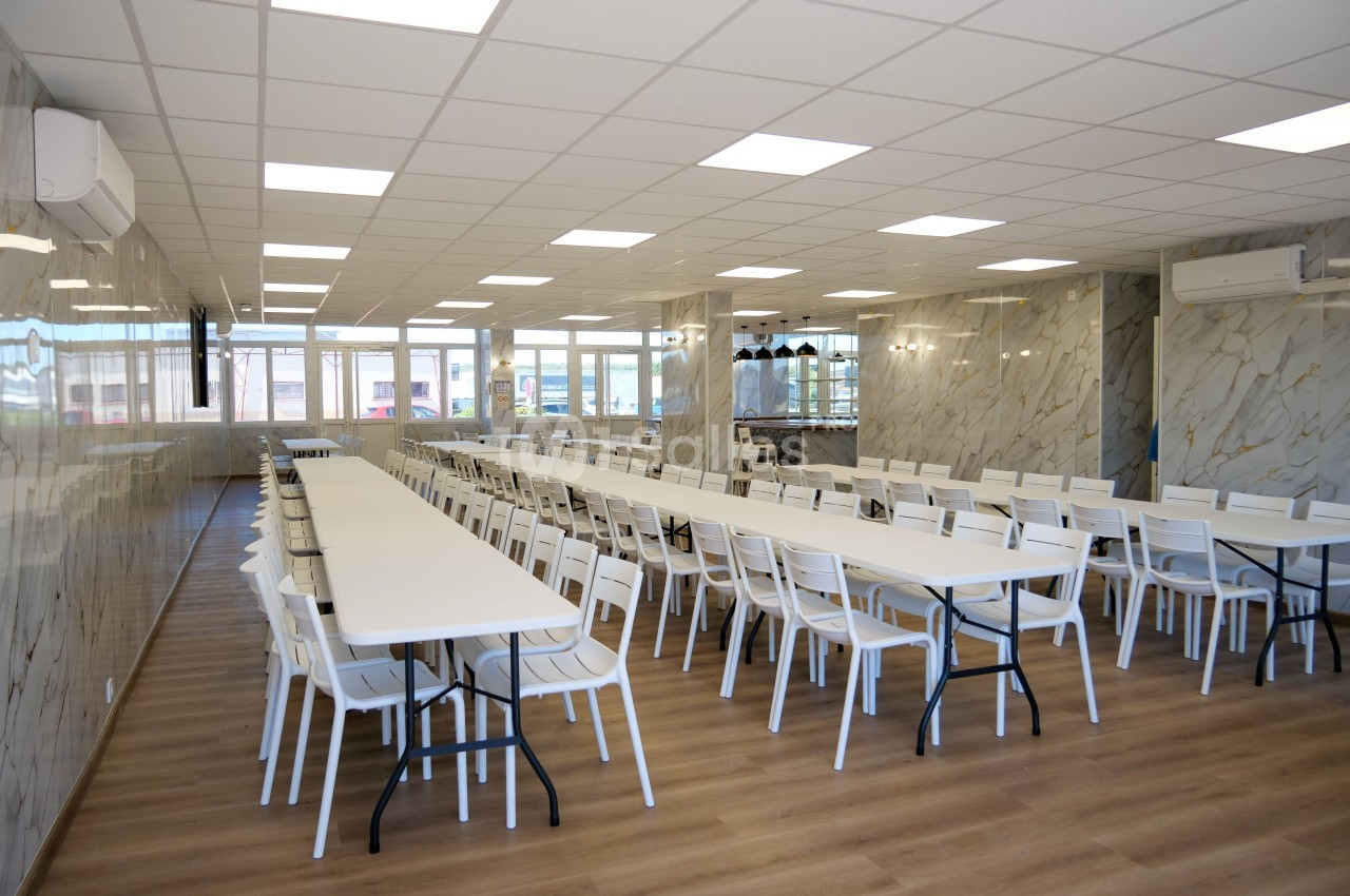Salle spacieuse avec de longues tables blanches alignées et des chaises, éclairée par des plafonniers et des fenêtres.