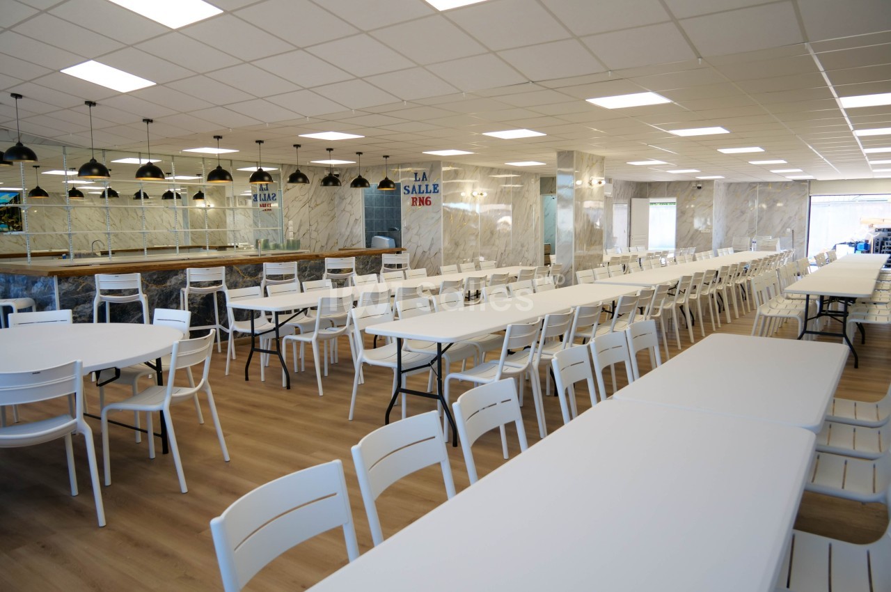 Salle à manger spacieuse avec des tables blanches alignées et des chaises, éclairée par des lampes suspendues.