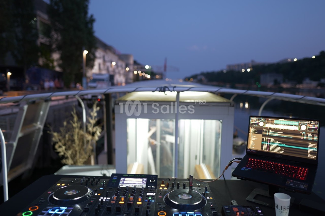 Poste de DJ avec platines et ordinateur portable sur un bateau, vue sur un canal urbain au crépuscule.