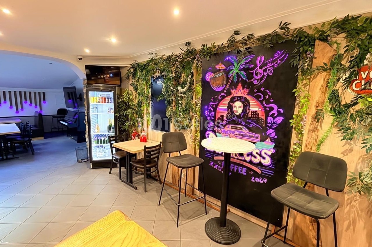 Intérieur d'un café avec décoration végétale, tableau mural coloré, tables et chaises modernes, et réfrigérateur en arrière…