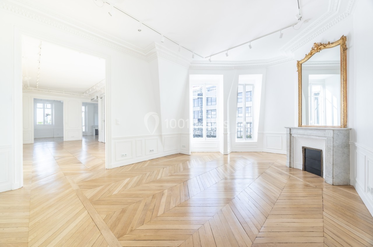 Intérieur lumineux avec parquet en chevrons, cheminée en marbre, miroir doré et grandes fenêtres donnant sur l'extérieur.
