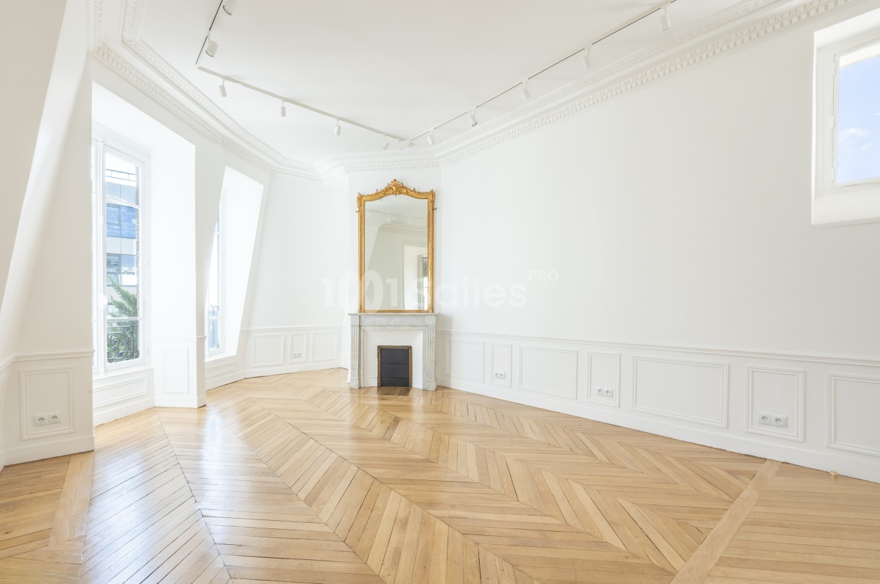 Salle lumineuse avec parquet en chevrons, cheminée surmontée d'un miroir doré et moulures au plafond.