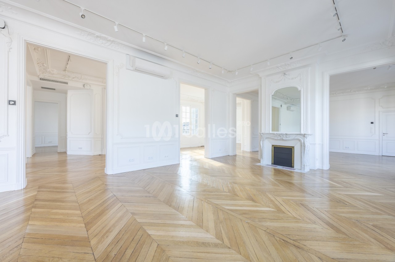 Intérieur lumineux d'un appartement vide avec parquet en chevrons, moulures blanches et cheminée avec miroir.