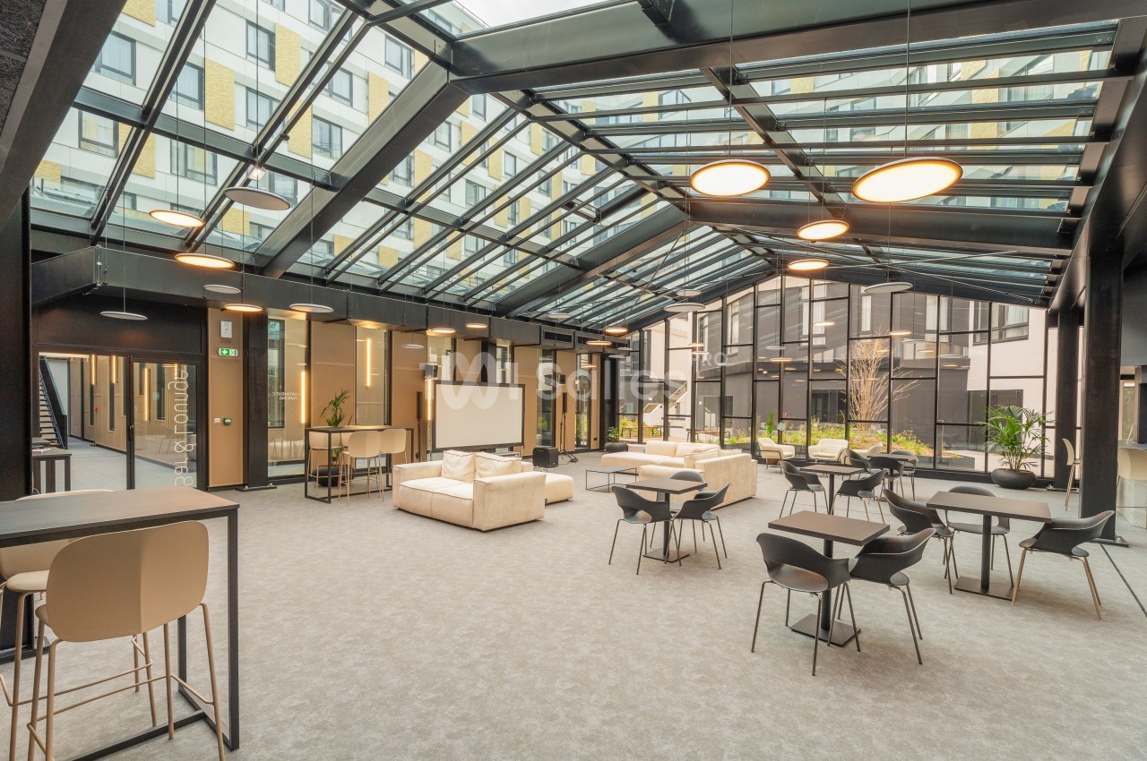 Espace intérieur lumineux avec verrière, mobilier moderne, tables, chaises et canapés, donnant sur une cour.