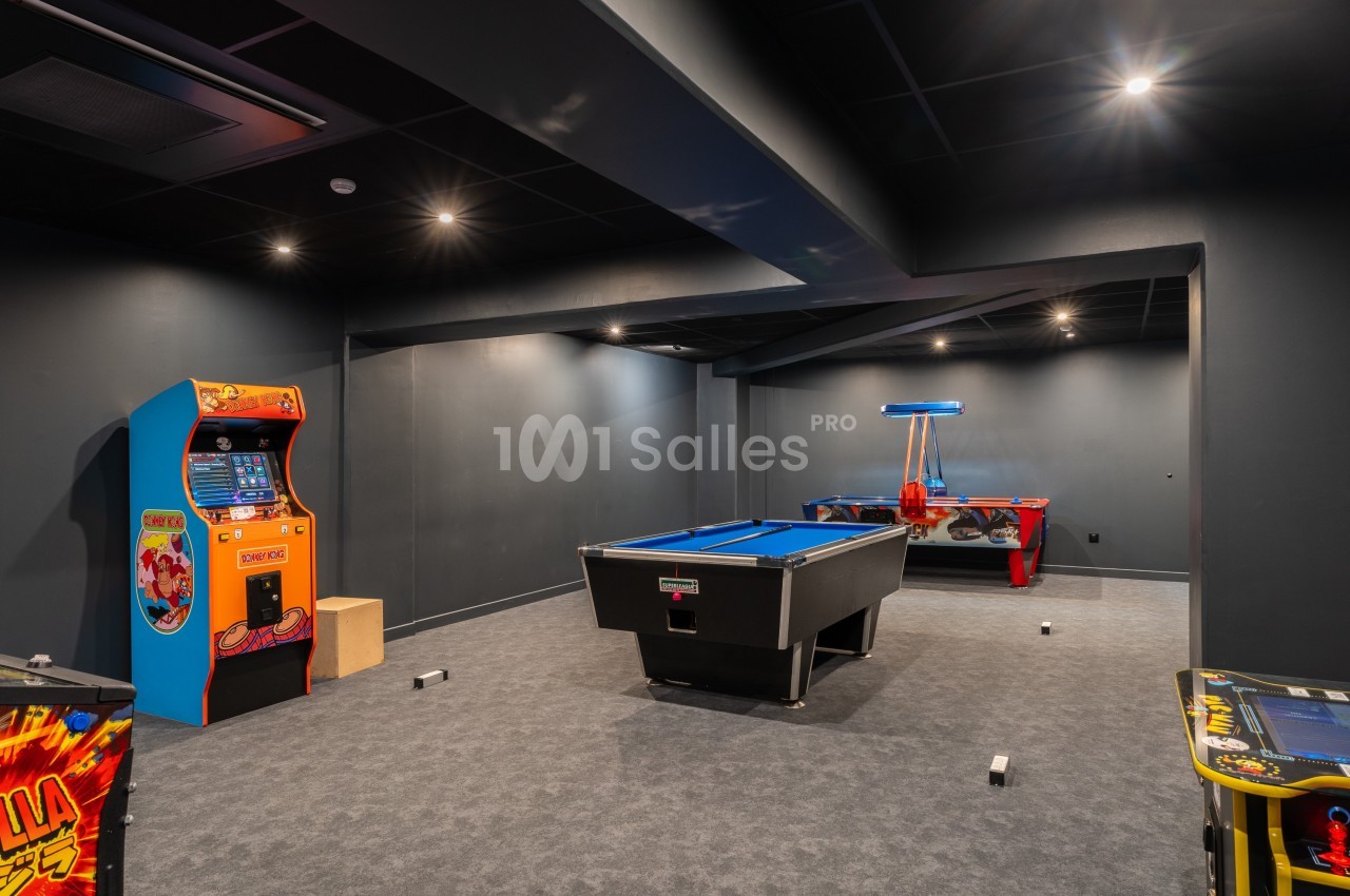 Salle de jeux avec un billard, une table d'air hockey et des bornes d'arcade dans un espace sombre et moderne.
